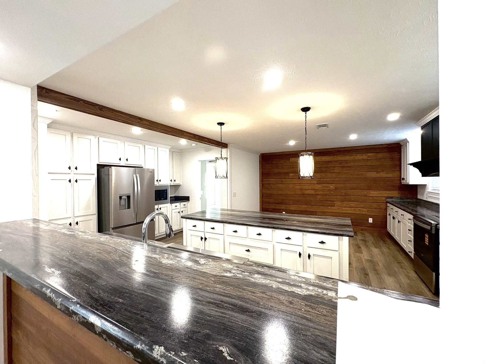 Modern kitchen featuring a long granite countertop with a faucet, sleek white cabinets, stainless steel appliances, wood flooring, and ambient pendant lighting.