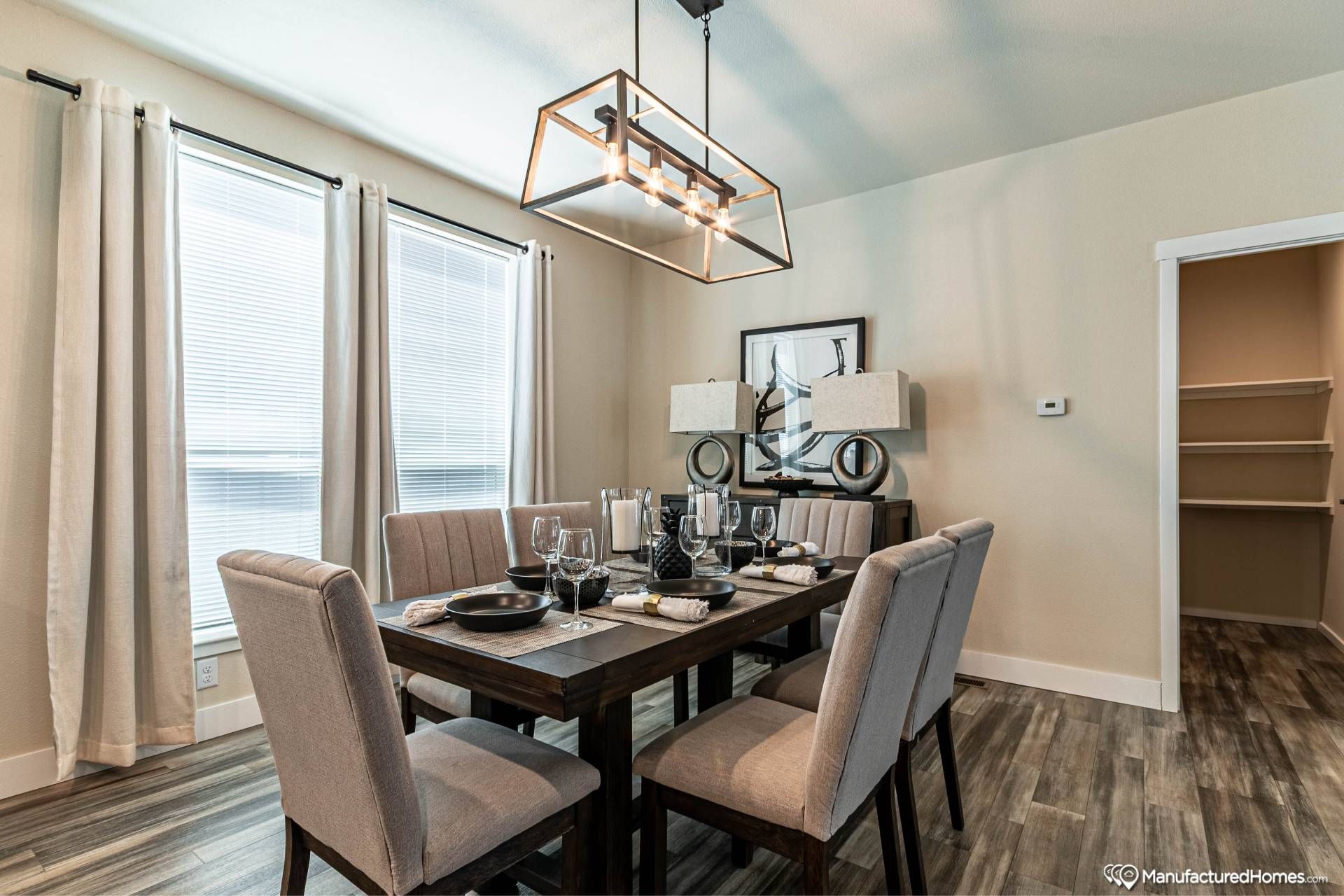 A modern dining room with a wooden table set for six, featuring elegant gray upholstered chairs. A geometric chandelier hangs overhead, and light streams in through tall windows with sheer curtains. Artwork and twin lamps adorn a sideboard, adding a contemporary touch. The room conveys a warm, inviting atmosphere.