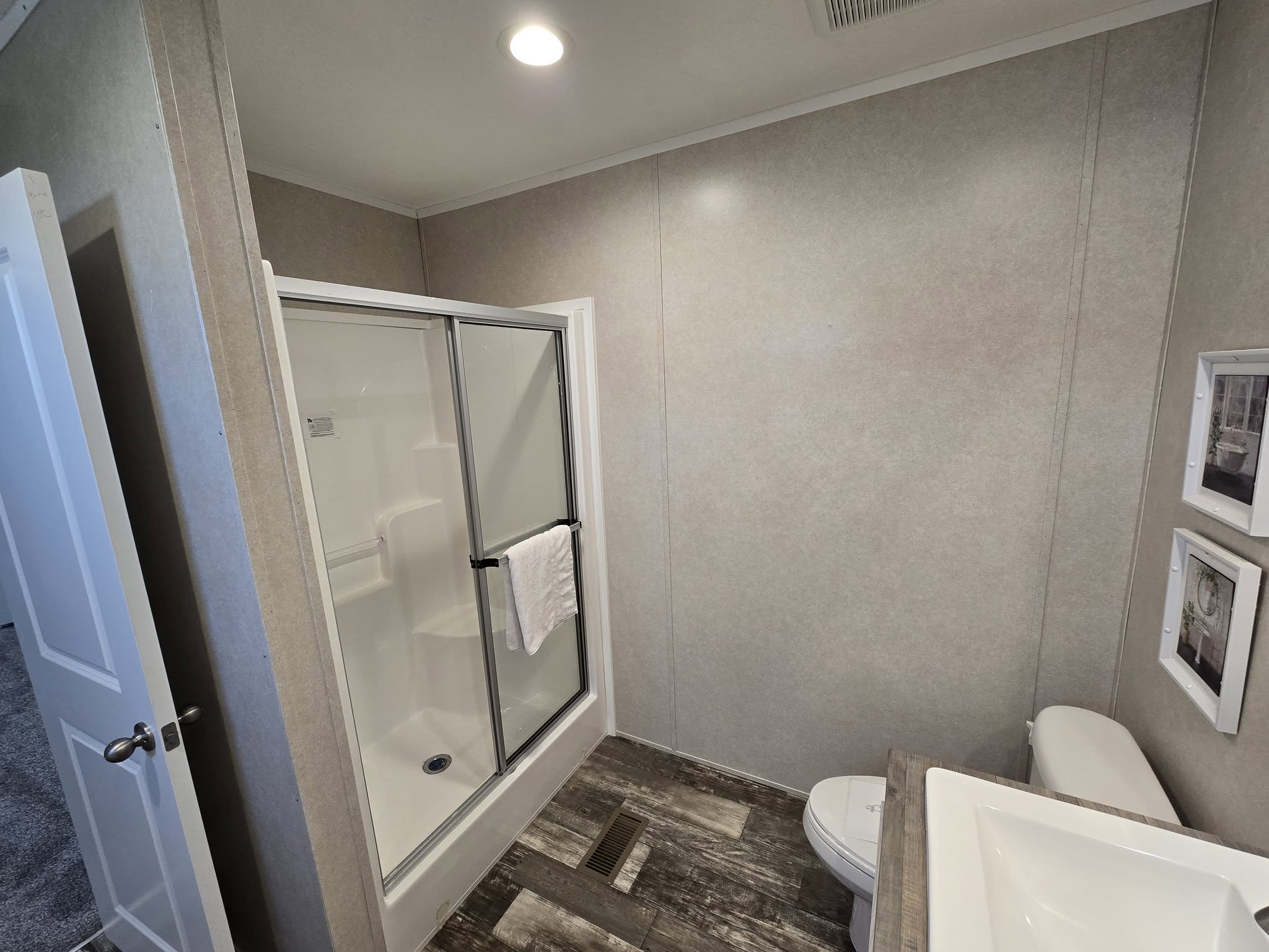 A modern bathroom features a shower with glass doors, a towel draped over, wall-mounted pictures, a toilet, and a sink, conveying a clean and minimal design.