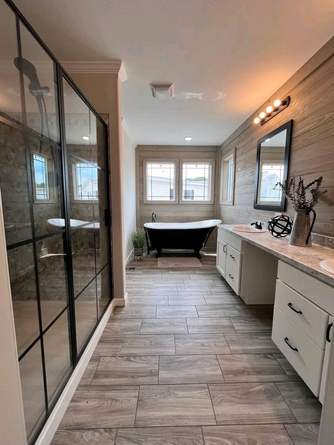 Modern bathroom with wood-tone floors and walls, featuring a black freestanding tub by three windows. A glass shower is on the left; vanity with mirror on the right. Bright, calming ambiance.