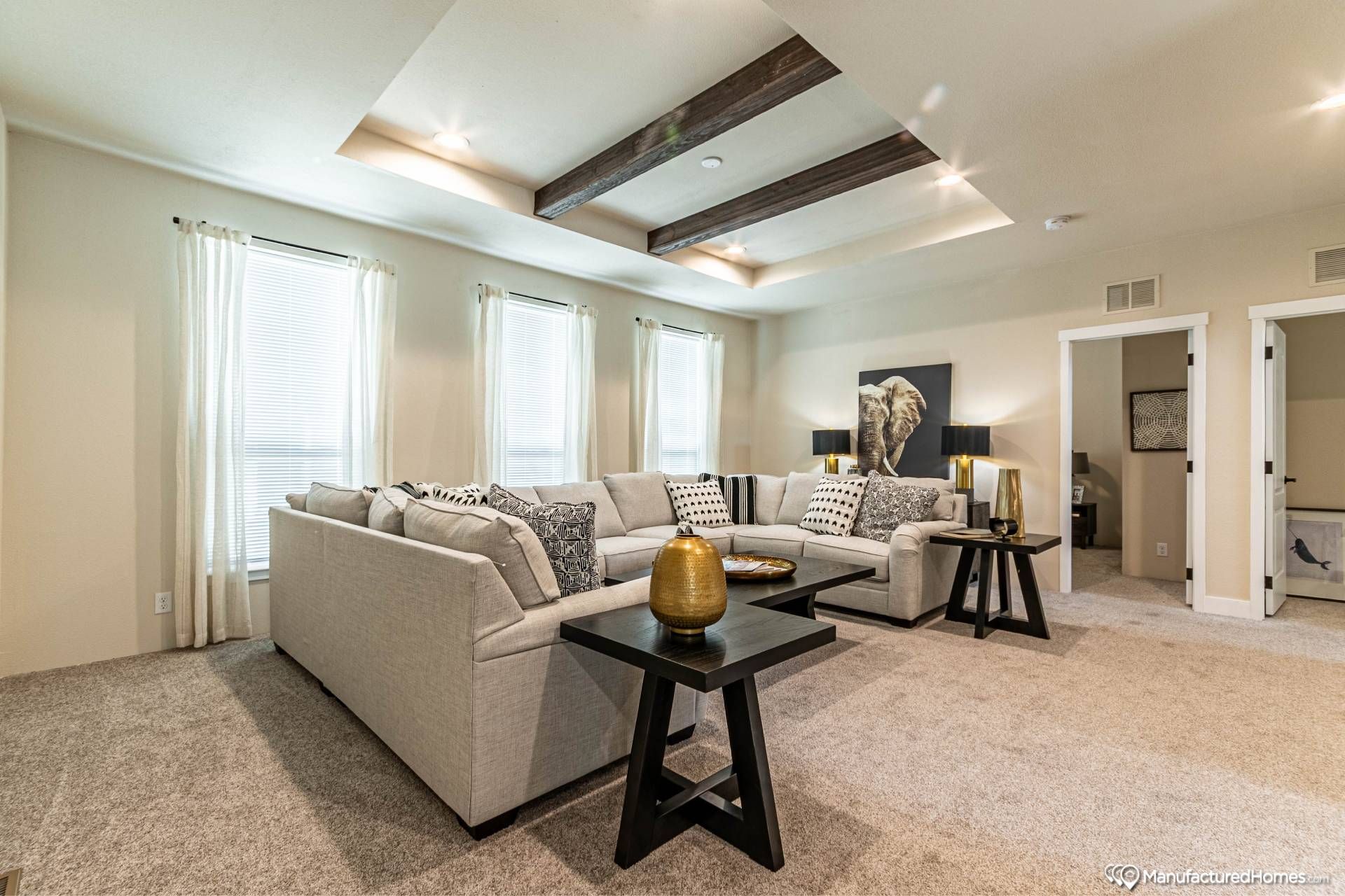 Spacious living room with beige sectional sofa, patterned cushions, and dark coffee tables. Large windows and ceiling beams add elegance and warmth.