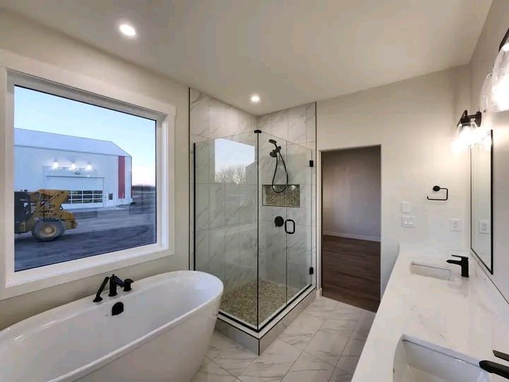Modern bathroom with a freestanding tub and glass shower. Large window displays an outdoor view. Bright lighting and sleek fixtures create a clean, tranquil ambiance.