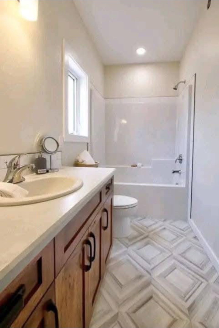Modern bathroom with wood cabinets, white countertop, and vessel sink on the left. A toilet and bathtub with shower are visible to the right. Bright lighting.