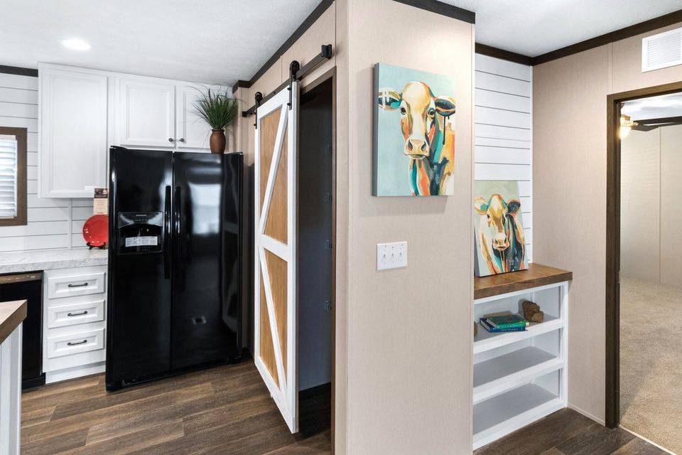 Modern kitchen with rustic barn door and black fridge. Art of a colorful cow hangs above white shelves, adding a playful touch to the room.