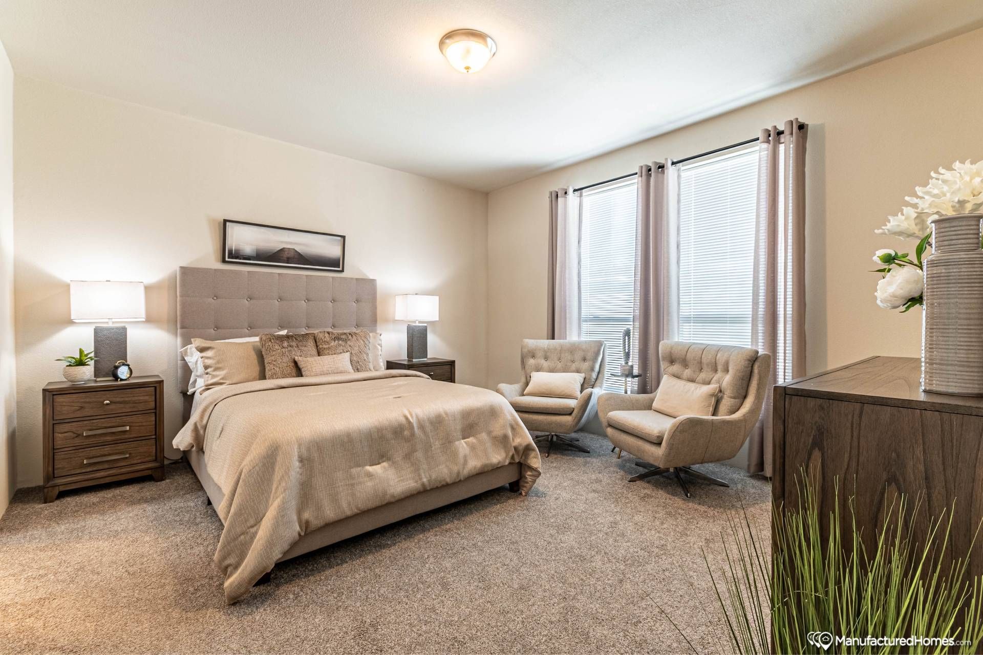 A cozy bedroom features a beige bed with plush pillows, flanked by wooden nightstands and lamps. Two gray armchairs sit near large windows with soft curtains.