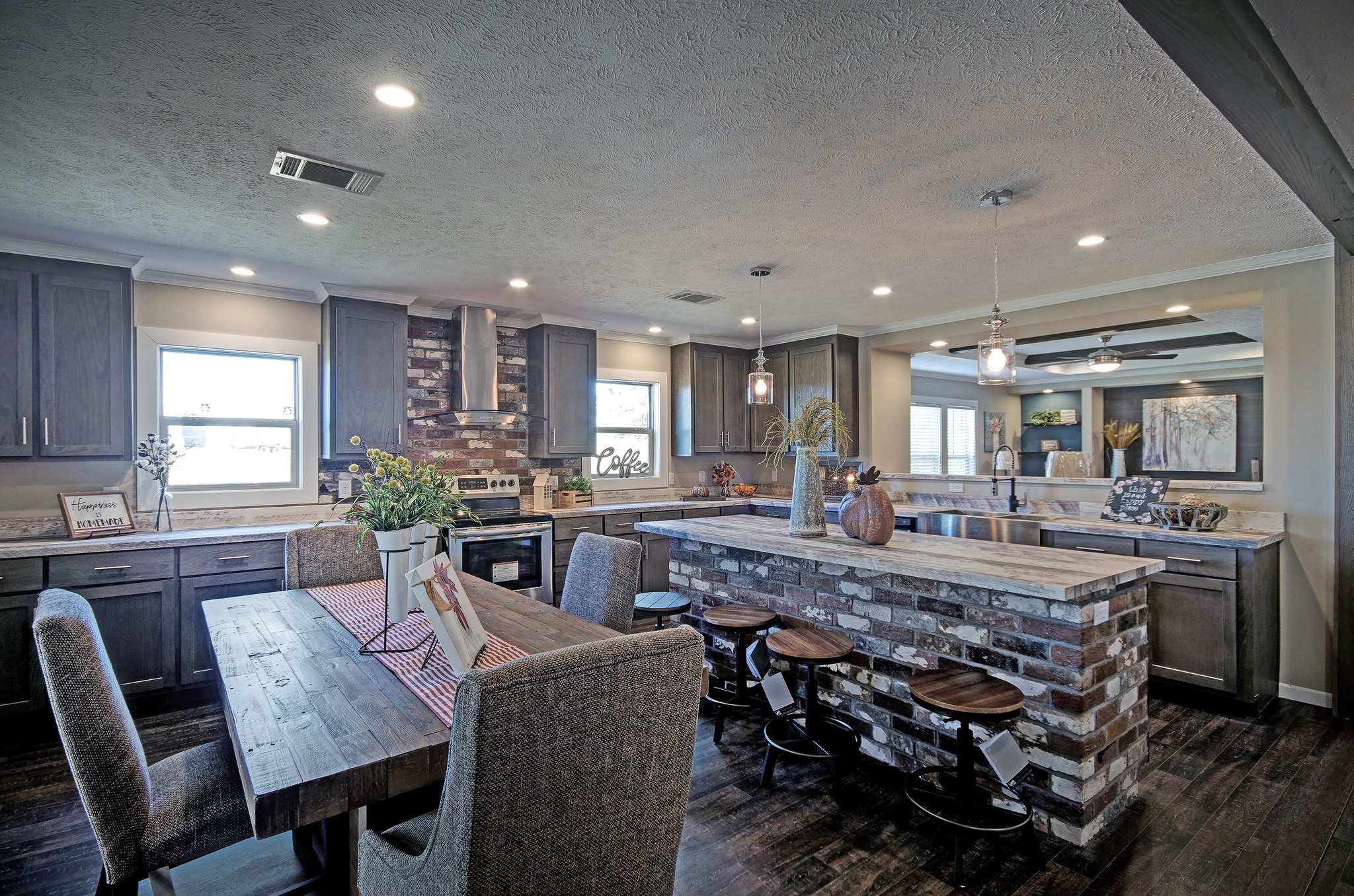 Modern kitchen with dark wood cabinets, brick accents, and an island with bar stools. A large dining table sits in the foreground. Warm, cozy ambiance.