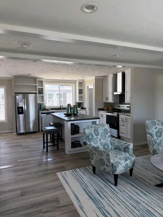 Modern open-plan kitchen and living area with wood floors. Features include a kitchen island, stainless steel appliances, and floral patterned chairs, creating a cozy and contemporary atmosphere.
