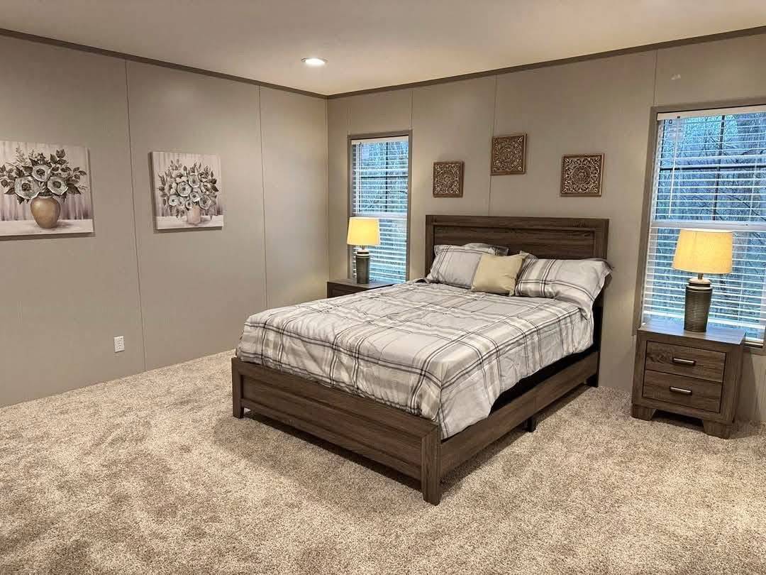 A cozy bedroom features a neatly made bed with plaid bedding, flanked by two wooden nightstands with lamps. Artworks and two windows add warmth. Neutral tones create a serene atmosphere.