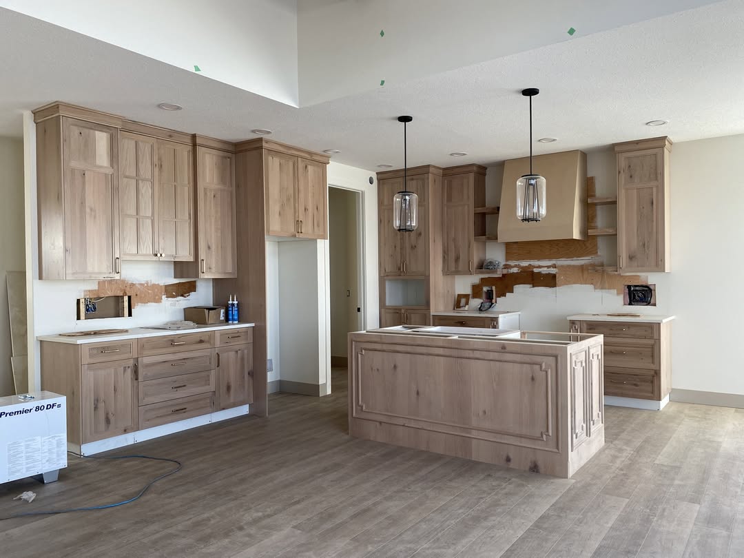 Modern kitchen under construction with light wood cabinets, an island, and pendant lights. The room has a calm, unfinished feel with exposed flooring.