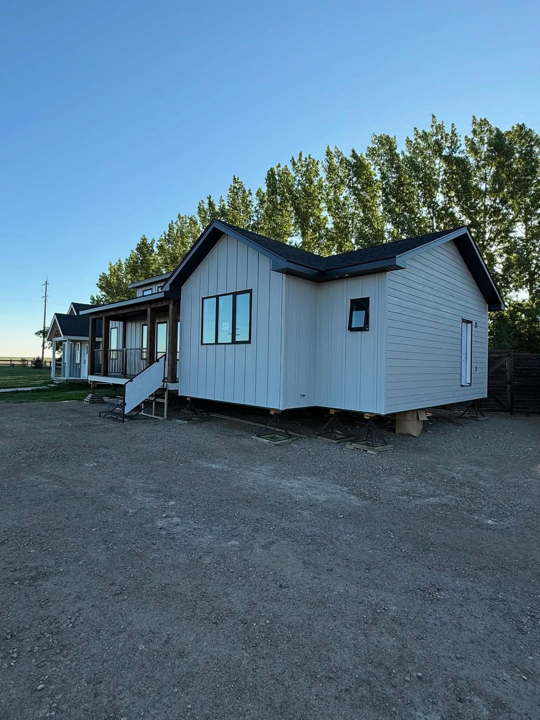 A small, light gray modular house sits elevated on blocks with a wooden porch and stairs, set against a backdrop of green trees and a clear blue sky.
