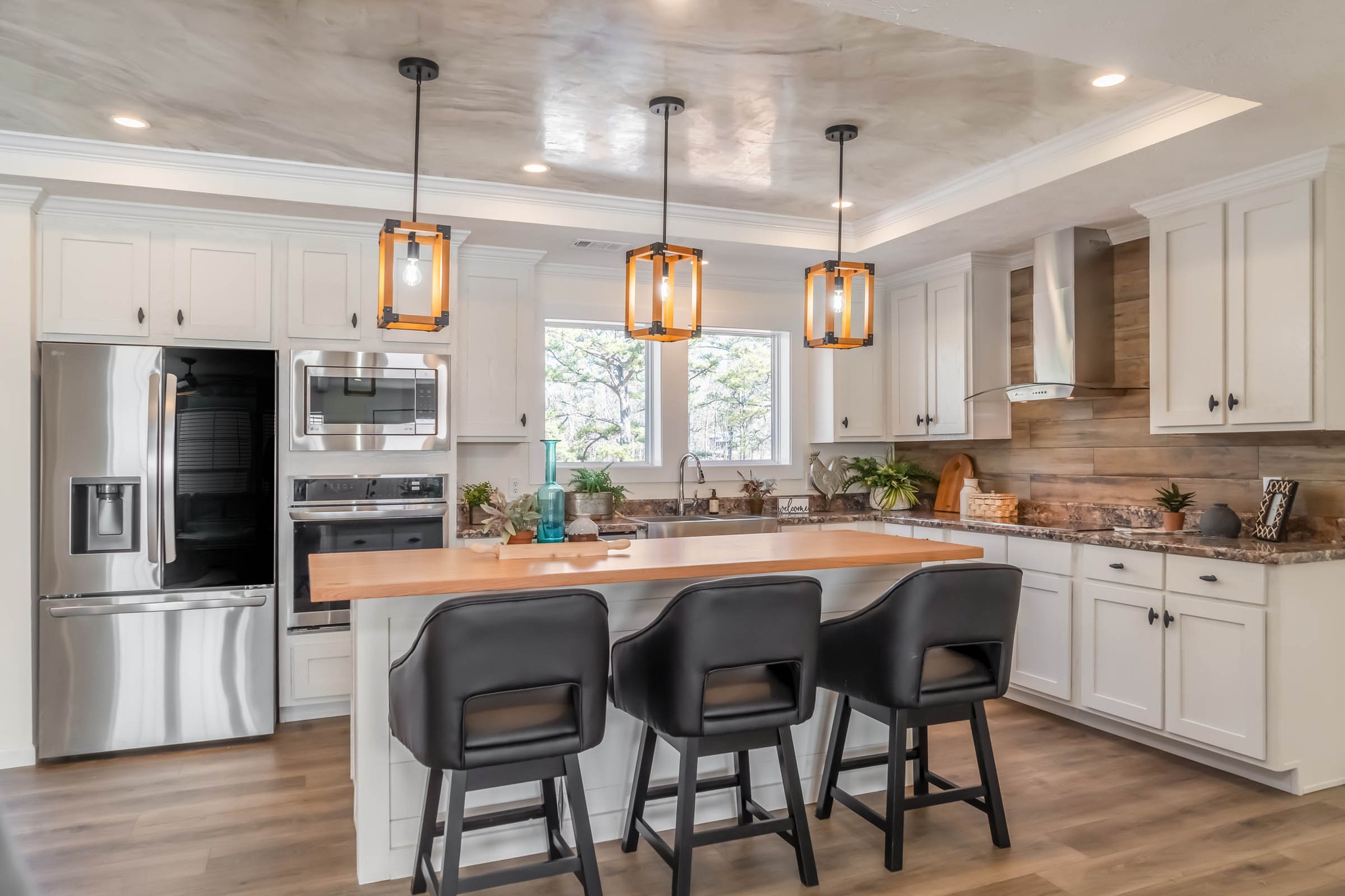 Modern kitchen with white cabinets, stainless steel appliances, and a wooden island with three black chairs. Pendant lights and plants add warmth.
