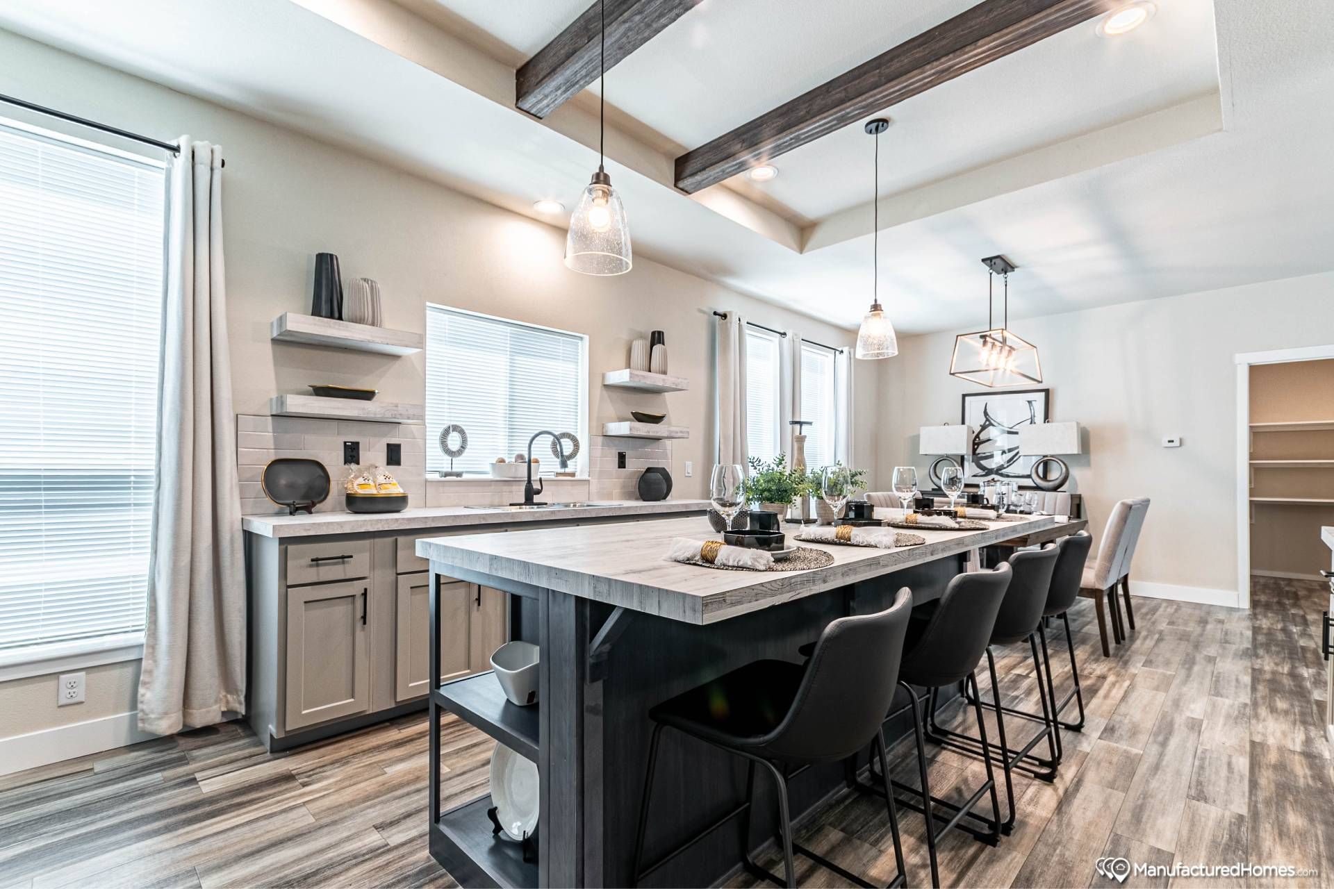 Modern kitchen with a long wooden island, six stools, pendant lights, and elegant decor. Bright and airy, with light wooden floors and natural light.