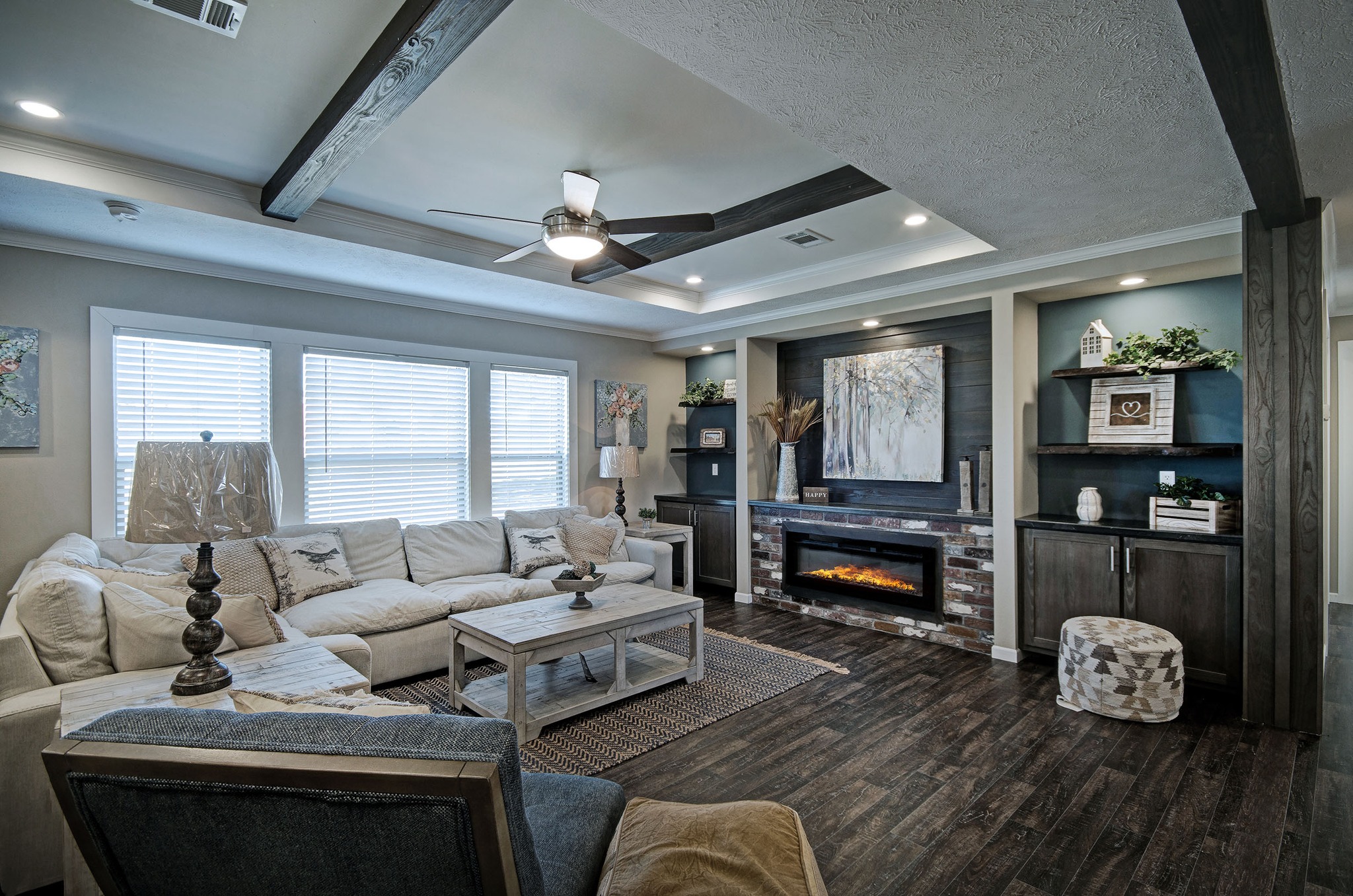 Cozy living room with a white sectional sofa and rustic decor. Features a modern electric fireplace, wooden beams, dark wood flooring, and a ceiling fan.