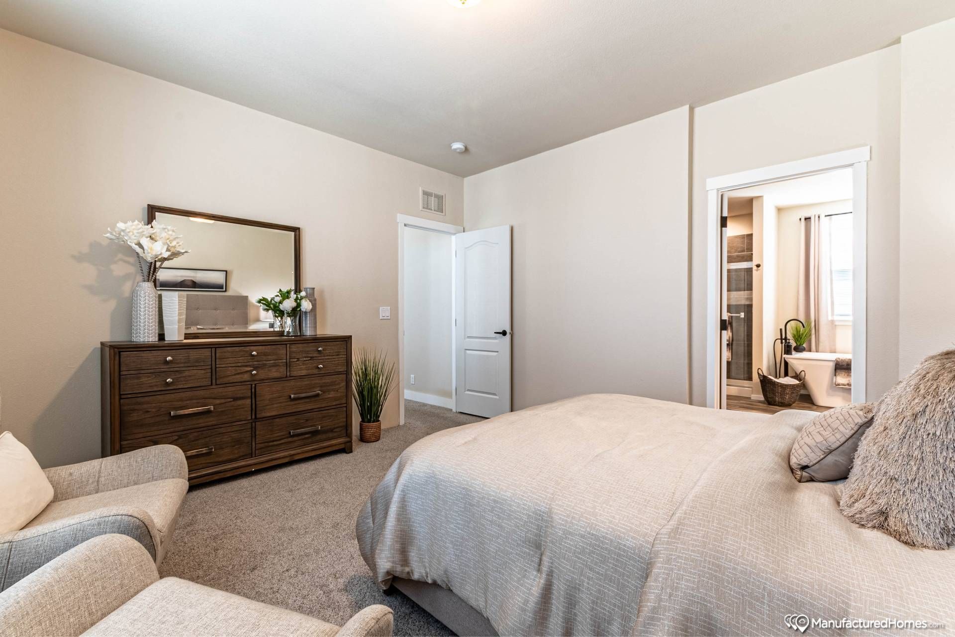 Modern bedroom with a beige theme, featuring a cozy bed with plush pillows, a wooden dresser with decor, and an open door leading to a bathroom.