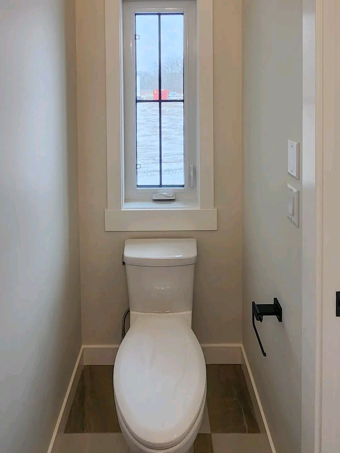 A narrow bathroom with a tall window above a white toilet. Light walls and a wooden floor create a simple, cozy feel. A toilet paper holder is on the right wall.