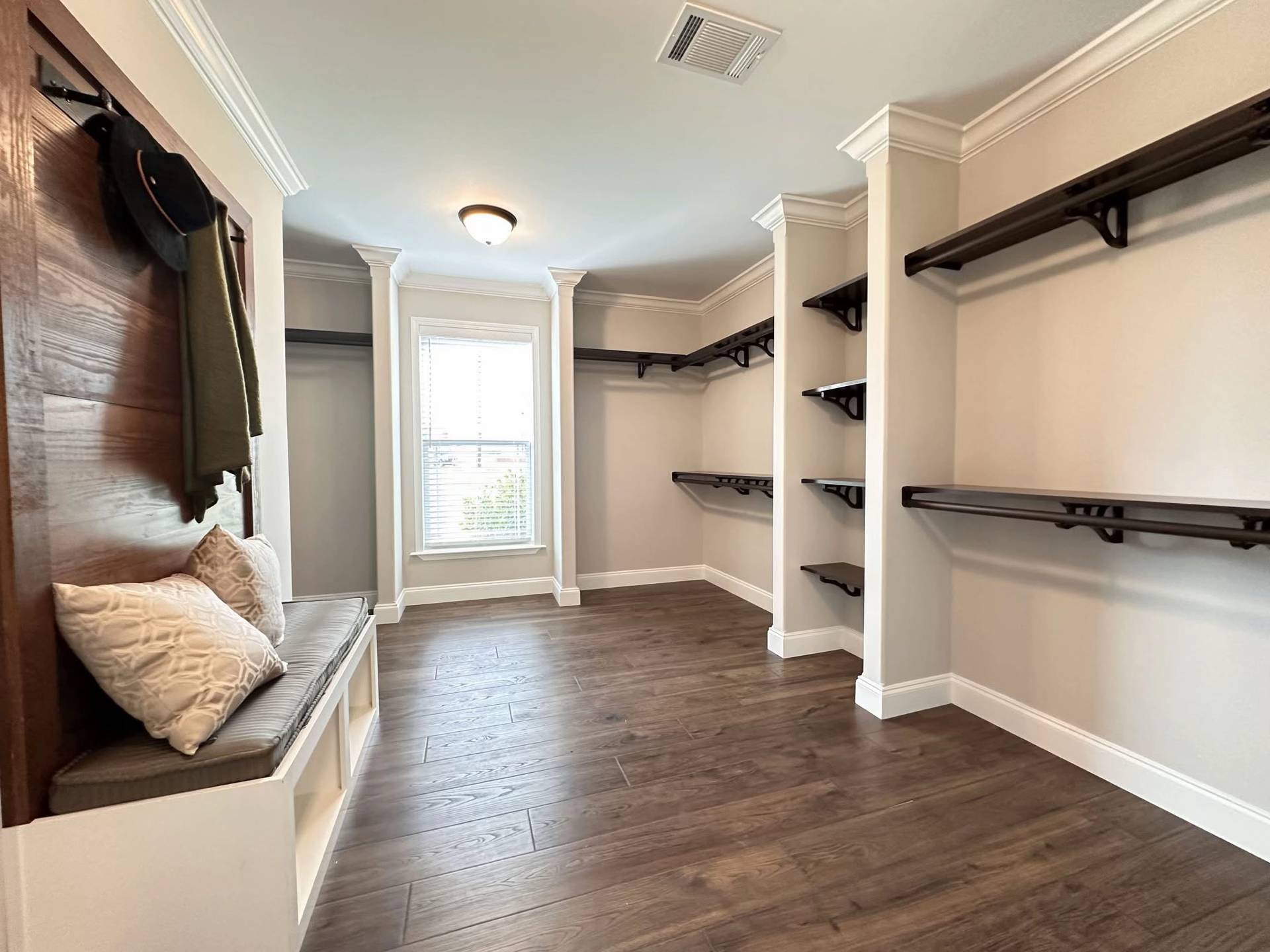 Spacious, well-lit walk-in closet with white walls and dark wooden shelves. A cushioned bench with pillows is on the left, under a hat and scarf.