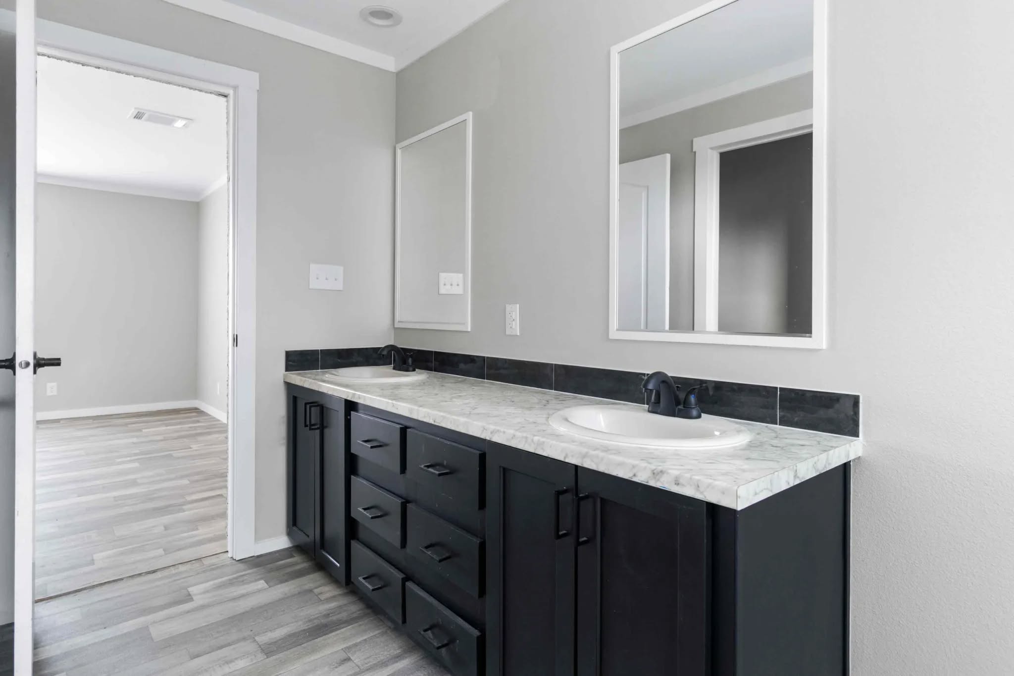 Modern bathroom with a marble countertop, black cabinets, and a single sink. A large mirror and open doorway lead to a room with wooden flooring.