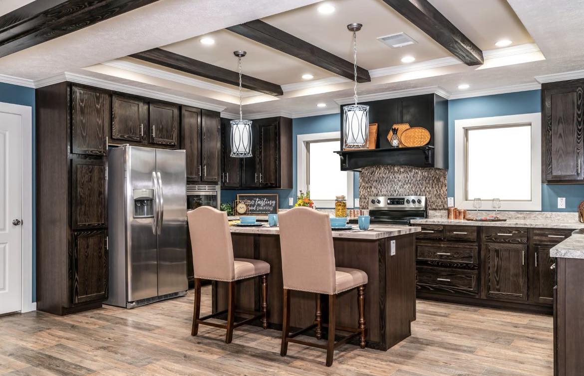 Modern kitchen with dark wood cabinets, stainless steel fridge, and island with beige chairs. Blue walls and ceiling beams create a warm, elegant feel.