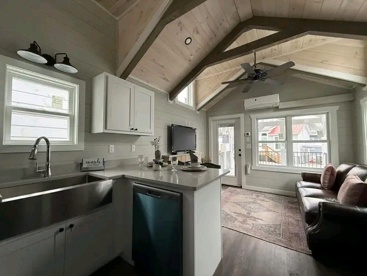 Cozy living space with vaulted wood ceiling, large windows, and a leather sofa. Modern kitchen with white cabinets and farmhouse sink. Airy and inviting.
