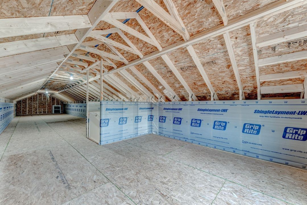 Expansive unfinished attic with exposed wooden beams and plywood flooring. Walls partially covered with insulation material labeled "Grip Rite."