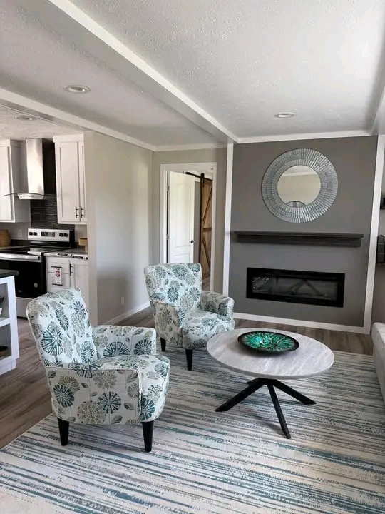Cozy living room with two floral armchairs facing a round coffee table. A modern fireplace and circular mirror create a stylish focal point. Bright and inviting.