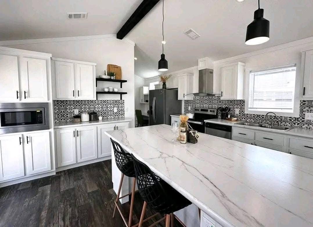 A modern kitchen with white cabinets, patterned black-and-white backsplash, and dark wood floor. A large marble island features two black chairs. Hanging pendant lights add a sleek touch.