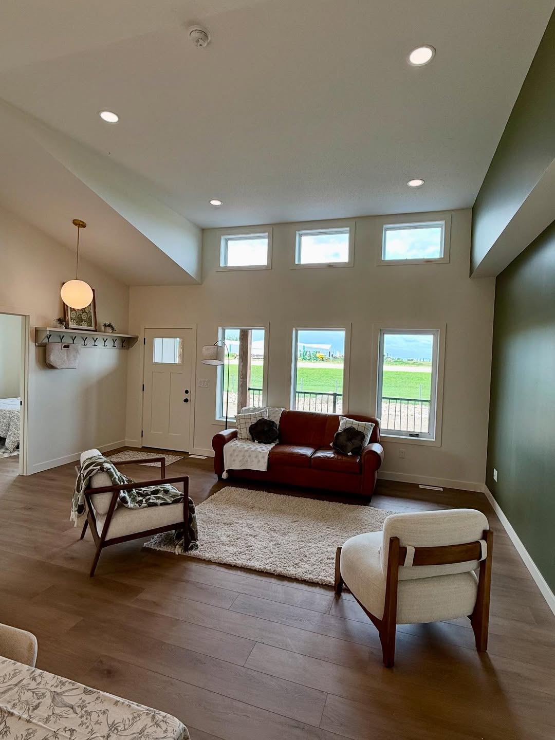 Spacious living room with a red sofa, two armchairs, green accent wall, and large windows showing a scenic view. Warm, cozy atmosphere.