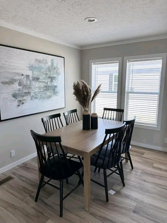 A minimalist dining room features a wooden table with six black chairs. A vase with dried pampas grass is centered on the table. Natural light streams through two windows, while a large abstract painting hangs on the wall, enhancing the serene ambiance.