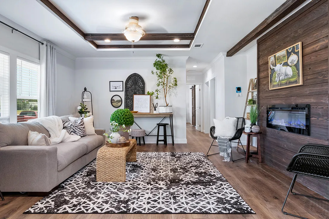 Cozy living room with a beige sofa, patterned rug, wooden floor, and a wall-mounted fireplace. Bright, airy space with plants and framed art, exuding warmth.