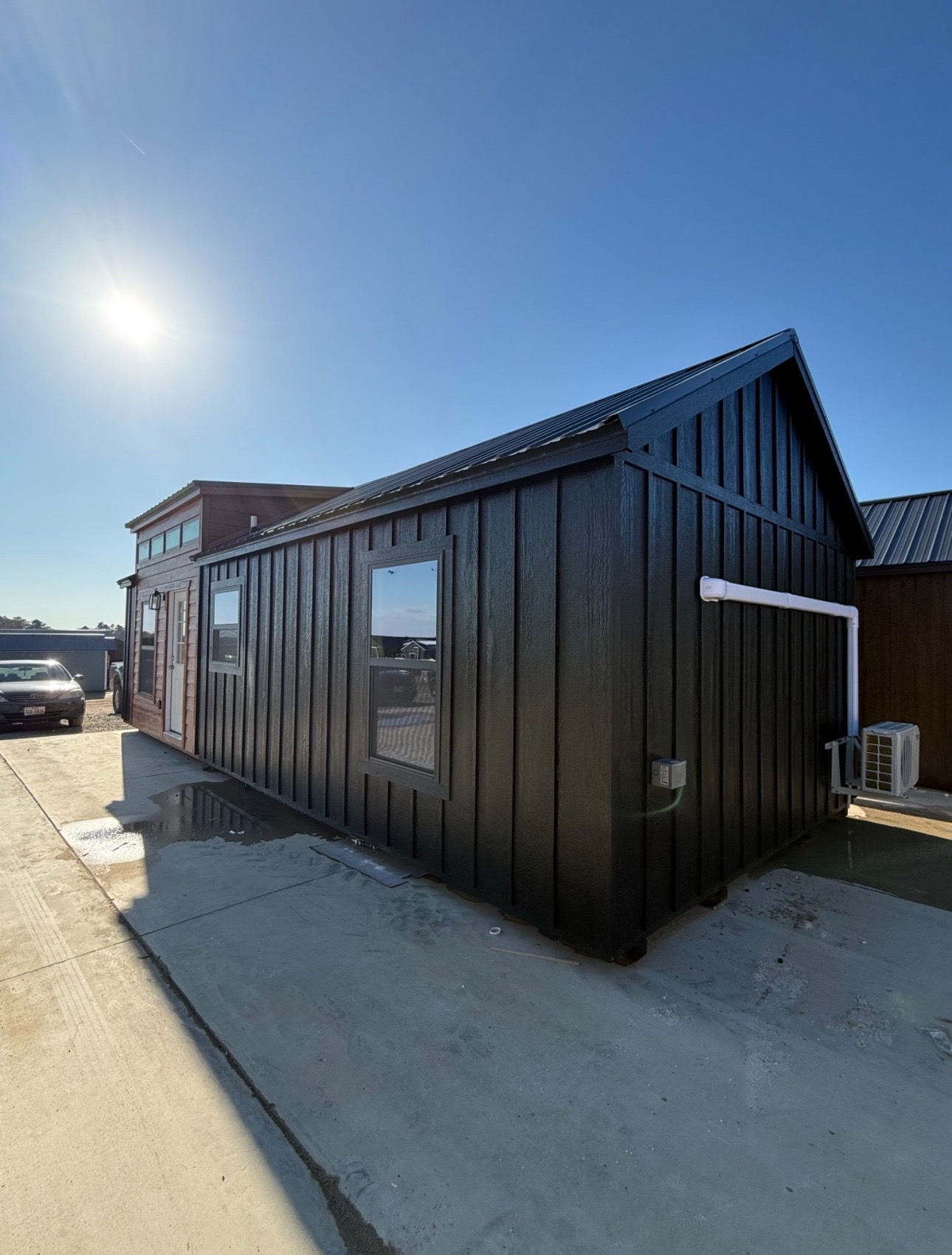 Small black house with a gabled roof and two windows, set on a concrete lot under a clear blue sky. The sun shines brightly, casting shadows.