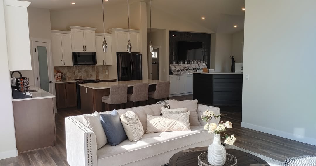 Modern living room with a beige sofa and decorative pillows, a round coffee table with flowers, open kitchen in the background with pendant lights, and a warm, inviting atmosphere.