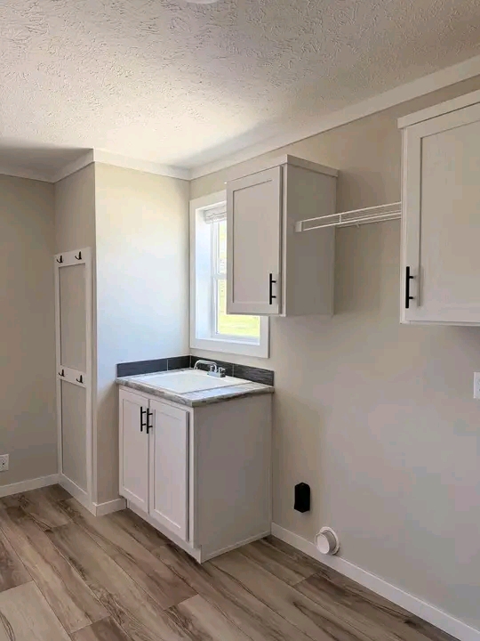 A small, bright utility room with light wood flooring. It features white cabinets, a small sink under a window, and a wire shelf. The tone is minimalist and clean.