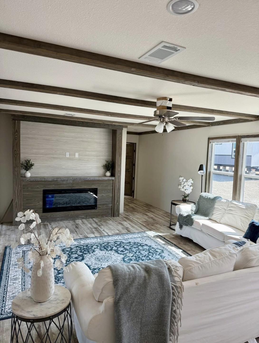 A modern living room with wooden beams, a ceiling fan, cream sofas, and a blue-patterned rug. Large windows let in light, creating a cozy, inviting atmosphere.