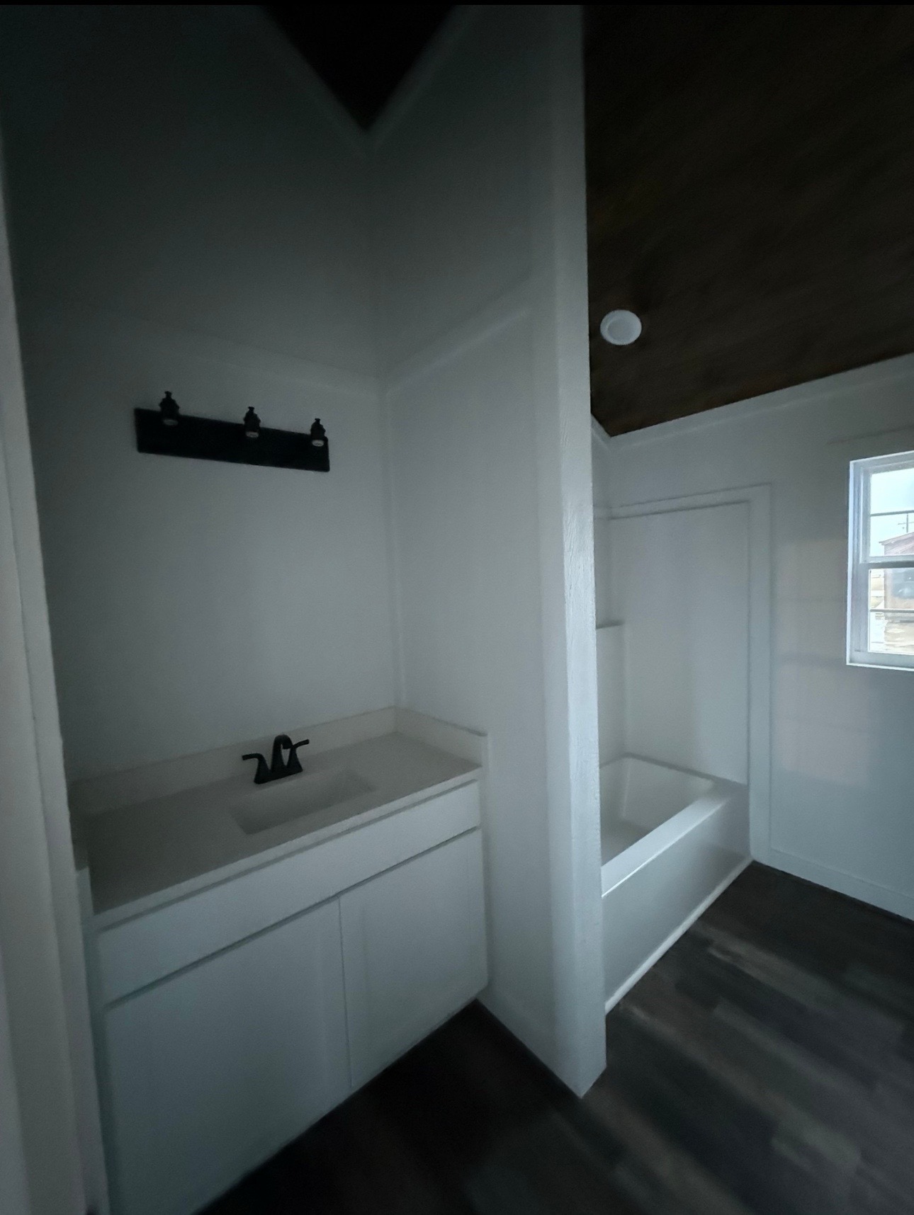Minimalist bathroom with dark wood floors, white walls, a white sink with a black faucet, recessed lighting, and a window above a bathtub.