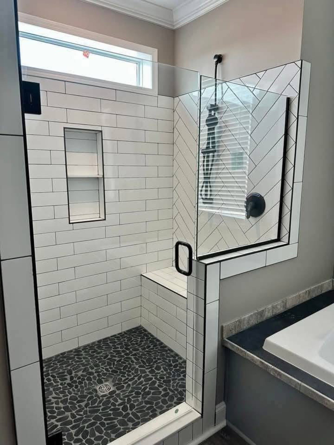Modern shower with white subway tiles and a black pebble floor. Glass door features a black handle. Light enters through a small window, creating an inviting and clean atmosphere.