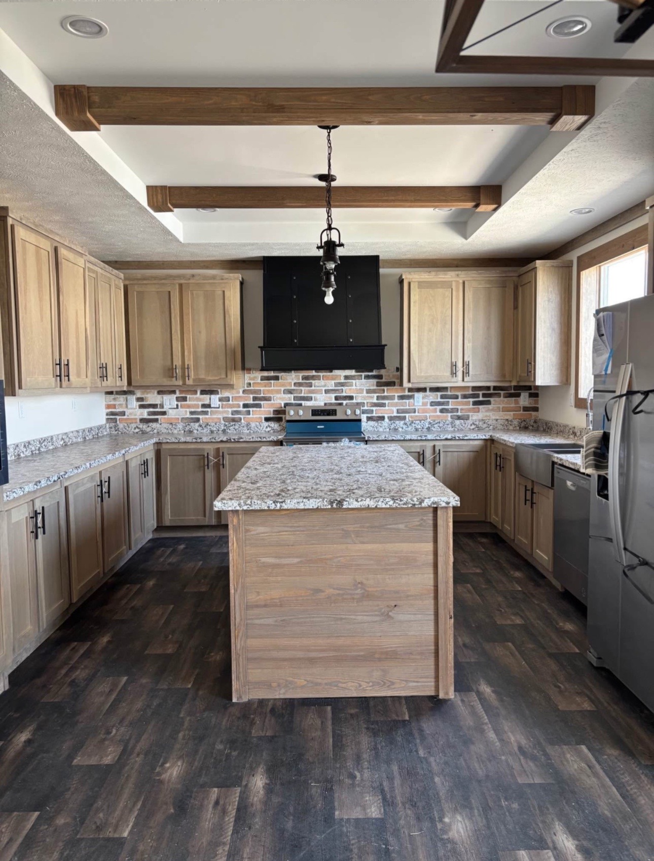 Spacious kitchen with wood cabinets, granite countertops, and a central island. Brick backsplash adds texture. Rustic beams and dark flooring create warmth.
