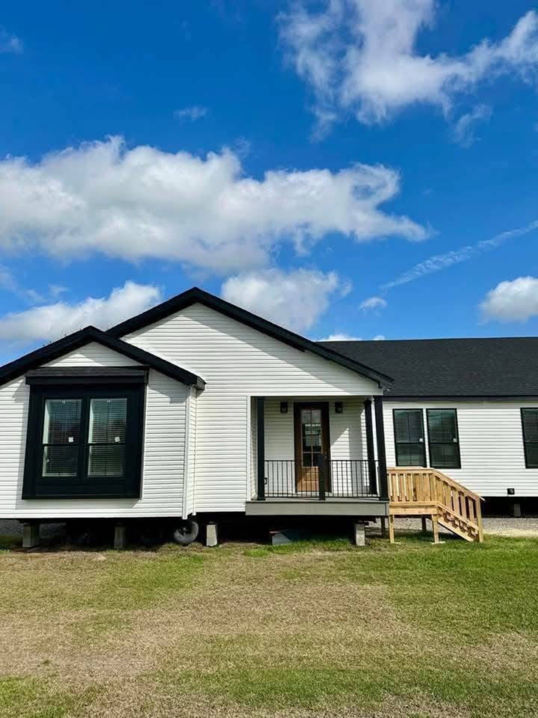 Single-story white house with black trim under a bright blue sky with fluffy clouds. The house has a small porch and a wooden ramp on the right.