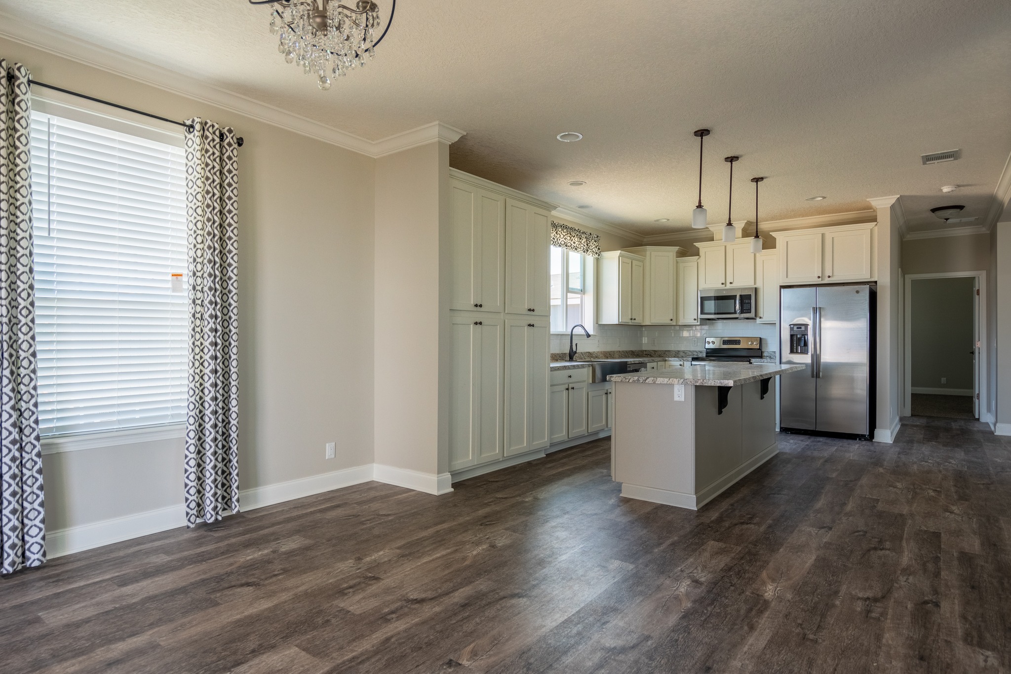 Spacious modern kitchen with dark wood floors, white cabinets, stainless steel appliances, and island. Sunlit room features patterned curtains and a chandelier.