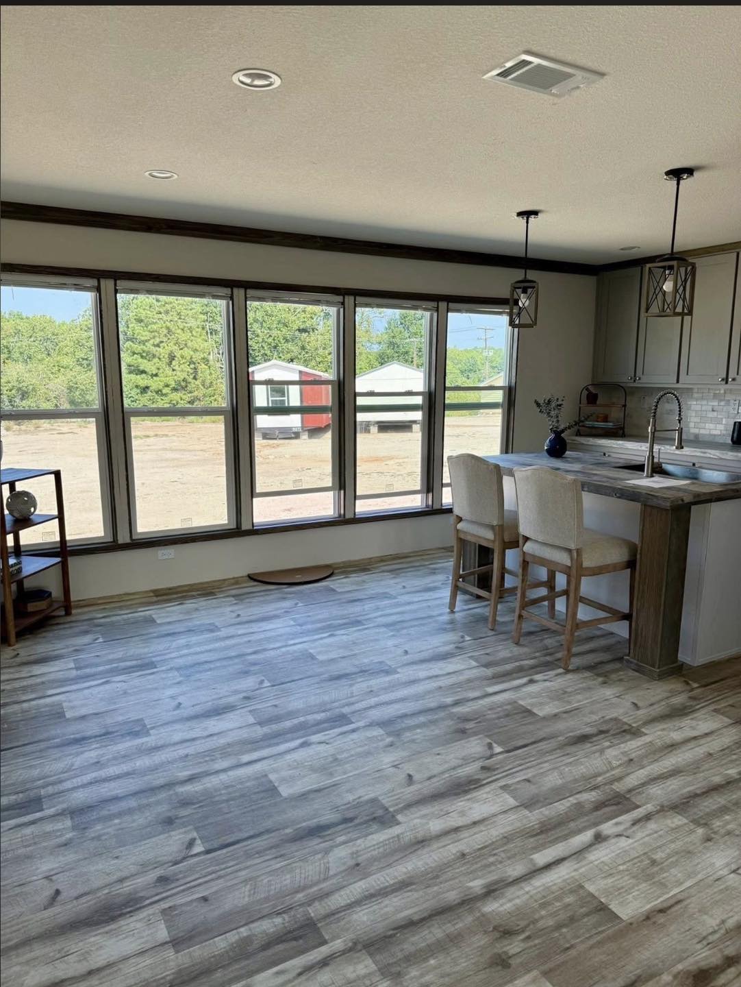 Modern kitchen with light wood flooring, a large window view of green trees and barns, and a white island with two chairs and hanging lantern lights. Cozy ambiance.