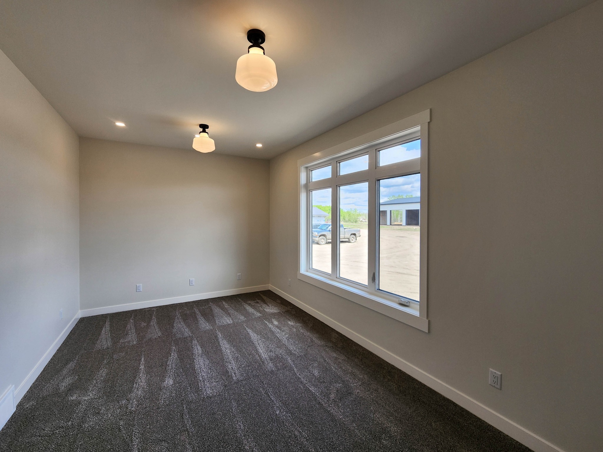A small, empty room with beige walls and a gray carpet. Two hanging light fixtures illuminate the space. A large window shows a sunny, outdoor view.