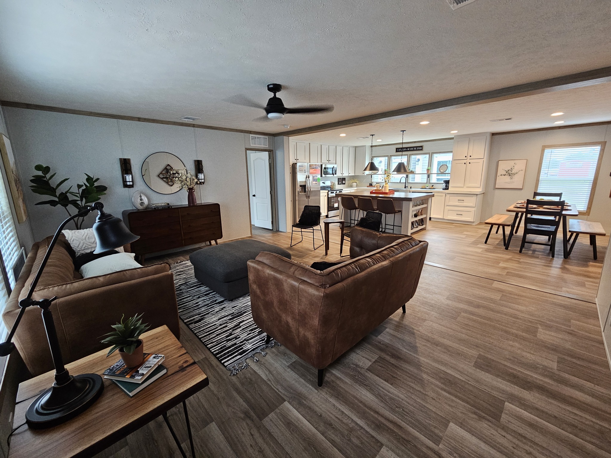 Modern open-concept living area with brown leather sofas, a kitchen island, and a wooden dining table. Warm lighting and decor create a cozy ambiance.