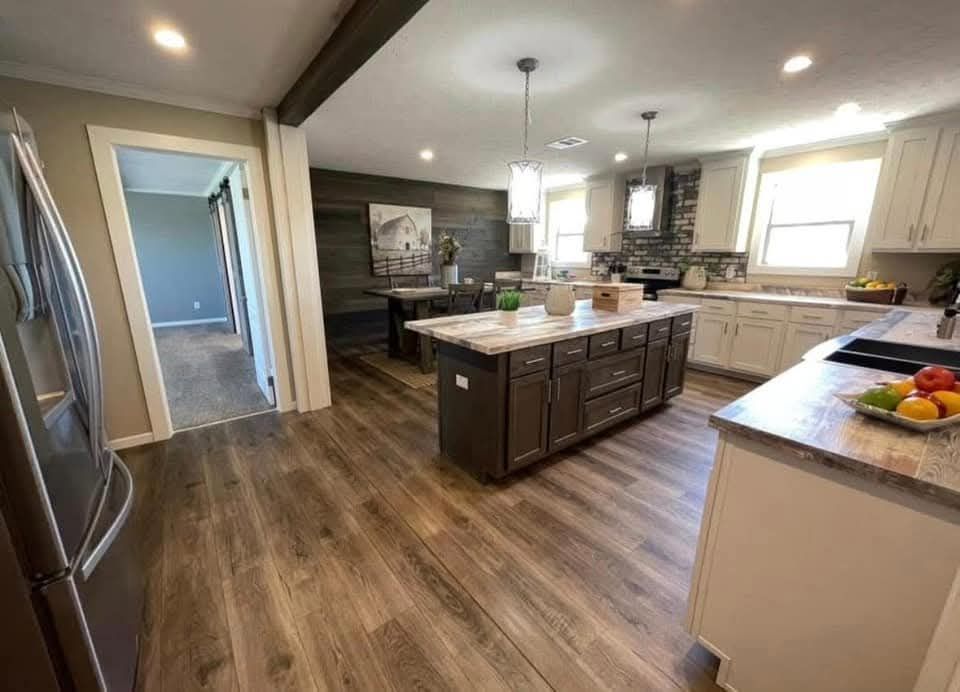 Spacious kitchen with wooden flooring, featuring a large island with dark cabinets, dual pendant lights, and a dining area. Bright, welcoming atmosphere.