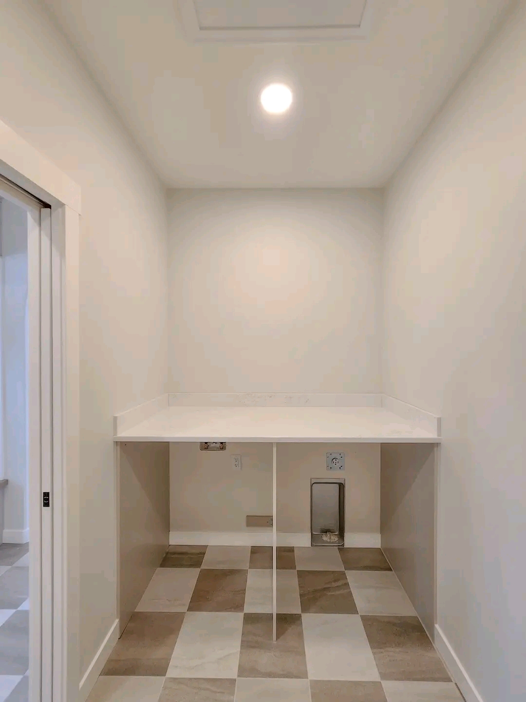 Laundry room with beige and white checkered tile flooring and a light beige wall. Overhead light brightly illuminates a countertop and utility connections.