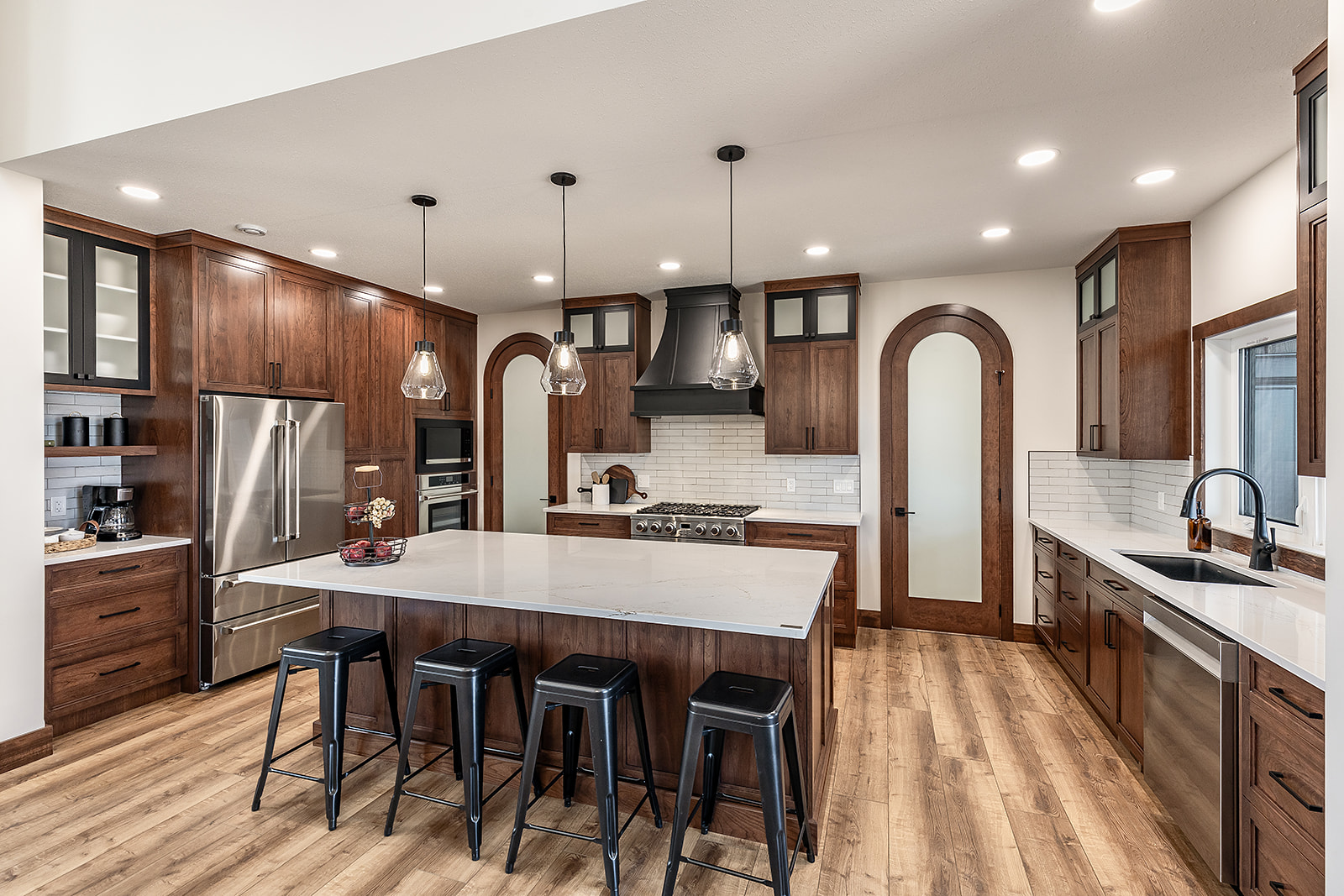 Modern kitchen with dark wood cabinets, white countertops, and stainless steel appliances. An island with black stools and pendant lights adds elegance.