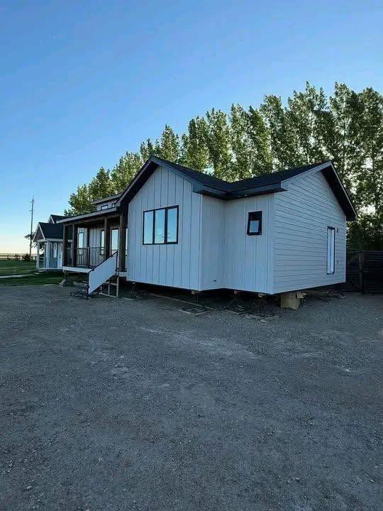 A small, white modular home with dark trim sits on a dirt lot, framed by trees and a clear sky. Its elevated design suggests mobility and modernity.