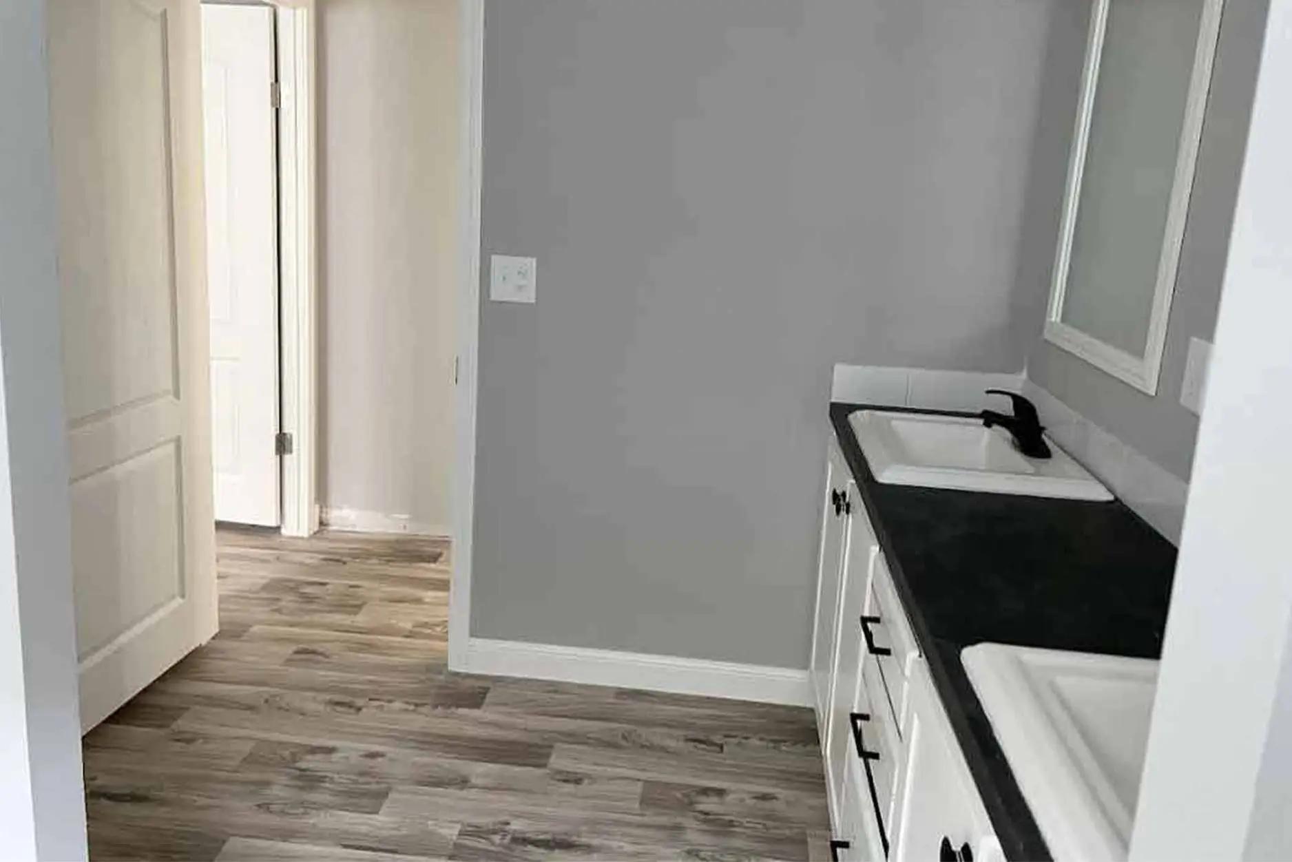 Modern bathroom with gray walls, wood flooring, and a dark countertop with two white sinks. An open door reveals an adjoining hallway. Cozy and clean.
