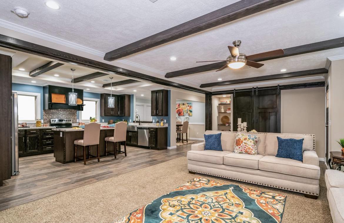 A spacious living room with beige sofas, colorful cushions, and a patterned rug. Exposed dark wood beams add warmth, while the open kitchen is seen in the background. A ceiling fan hangs overhead, enhancing the inviting atmosphere.