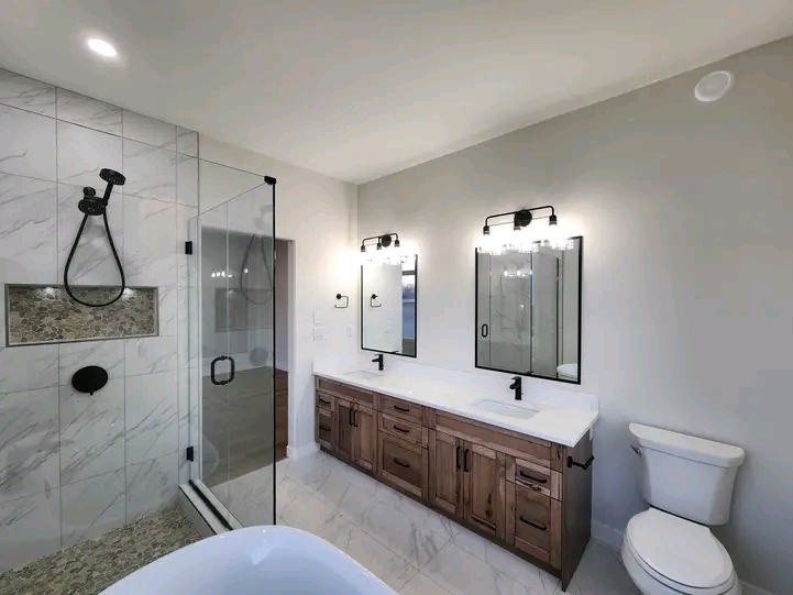 Modern bathroom with a glass shower, marble tiles, dual sink wooden vanity, and large mirrors. Bright lighting creates a clean, elegant feel.