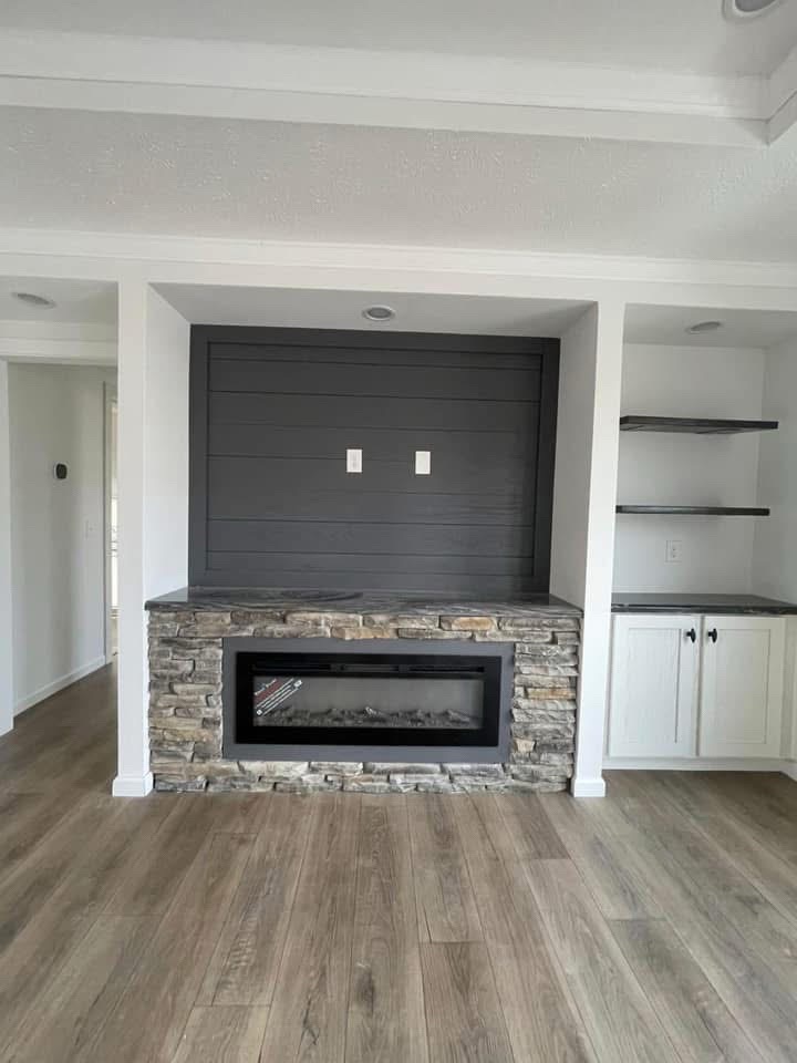 Living room interior with a stone fireplace and modern electric insert. Dark wood paneling and shelves on the right, light wood flooring, and a cozy ambiance.