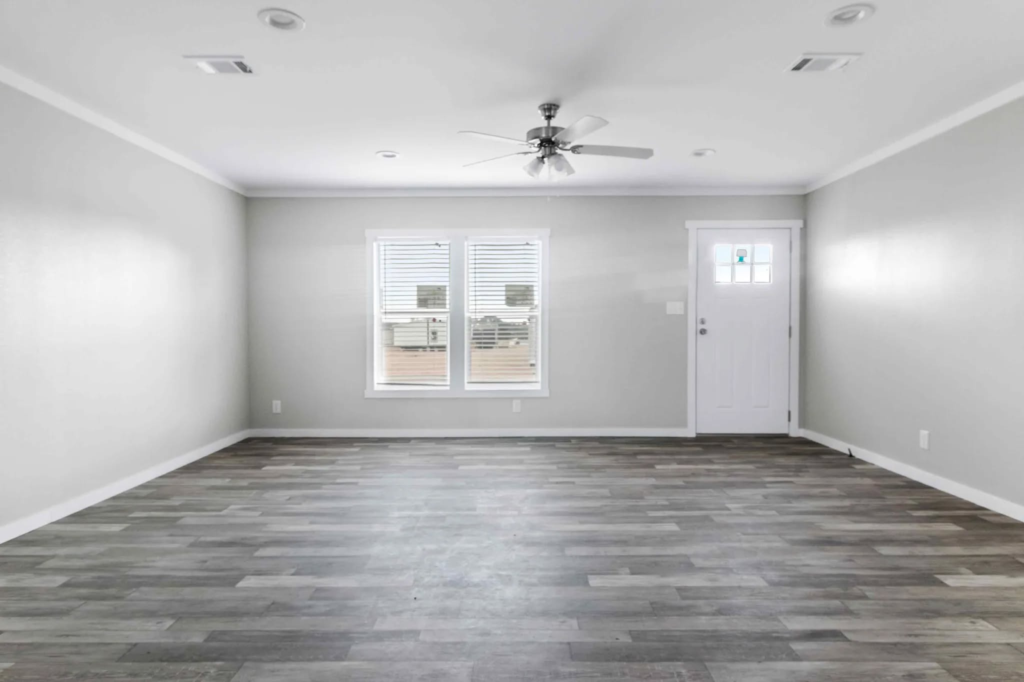 Empty room with light gray walls and wood flooring. Features two windows with blinds, a white door, and a ceiling fan, creating a spacious and neutral atmosphere.